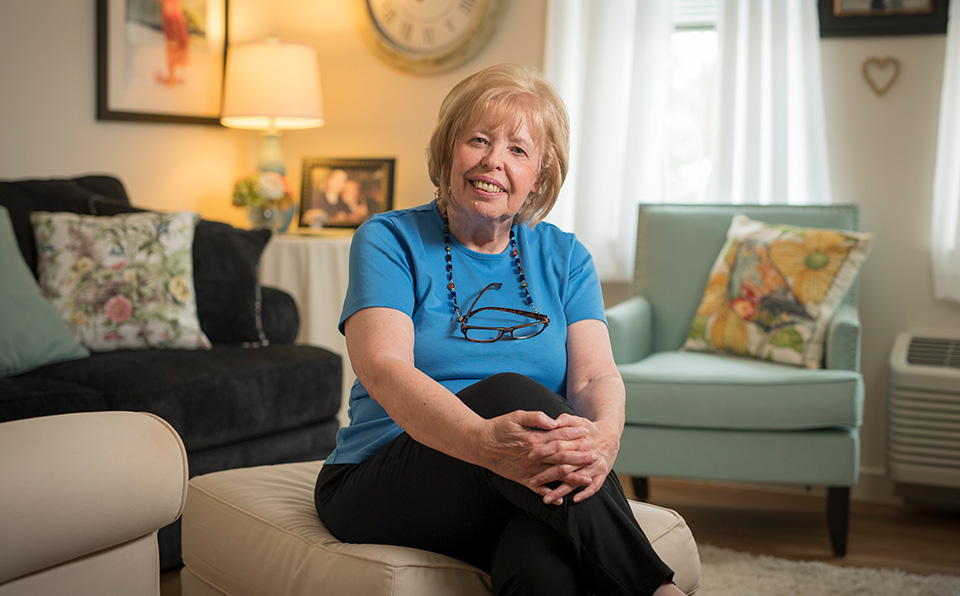 Older woman with friendly smile sits with arms and legs crossed looking at camera.