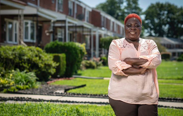 Smiling woman in front of a row of homes with beautiful lawns stands with her arms crossed.